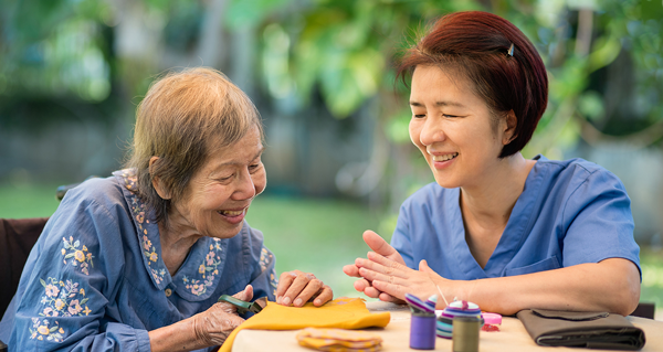 Duas mulheres da Ásia Oriental estão sentadas numa mesa fazendo artesanato juntas. Uma mulher é idosa e a outra é sua assistente de cuidados pessoais.