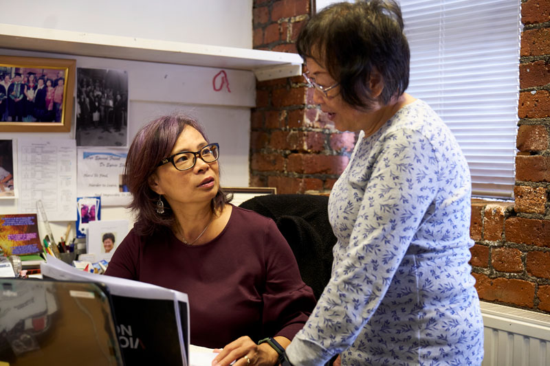 Una empleadora, una mujer de Asia Oriental sentada frente a su mesa, habla con otra empleadora, una mujer de Asia Oriental mayor que ella, que está de pie a su lado.