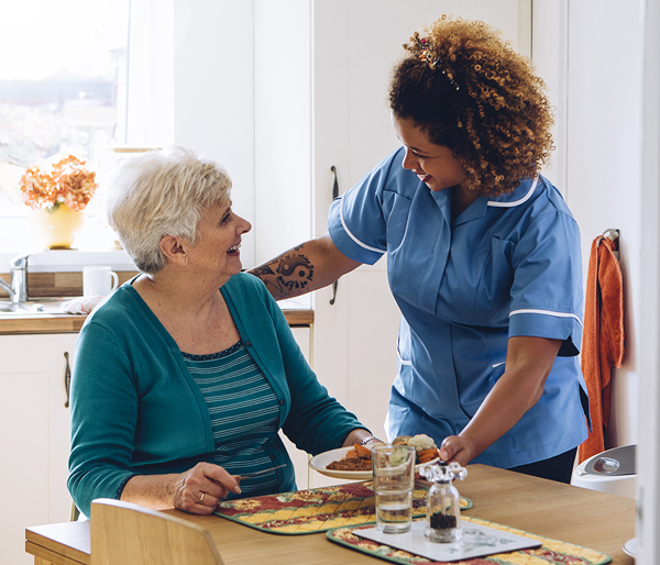 Uma mulher idosa branca está sentada à mesa da cozinha e sua assistente domiciliar, uma jovem negra com uma tatuagem, traz-lhe a comida.